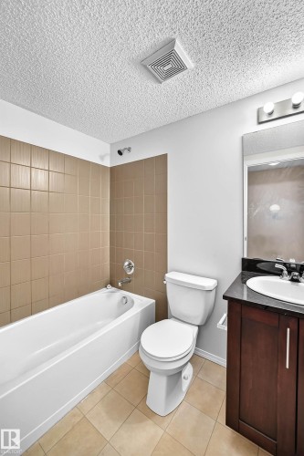 Bathroom featuring a white bathtub with chrome fixtures, brown tiled shower surround, light beige floor tiles, and a dark wood-finish vanity with a dark countertop and undermount sink - #404 12025 22 Av Sw, Edmonton, AB - Indoor Photo Showing Bathroom
