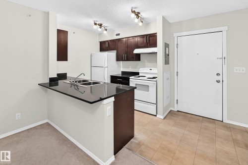 Kitchen featuring dark wood-finish cabinetry, black countertops, and a double basin sink - #404 12025 22 Av Sw, Edmonton, AB - Indoor Photo Showing Kitchen With Double Sink