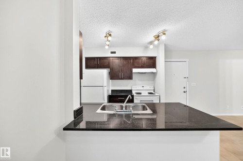 Kitchen featuring a dark countertop with an integrated double basin sink, dark wood-finish cabinetry, and white appliances - #404 12025 22 Av Sw, Edmonton, AB - Indoor Photo Showing Kitchen With Double Sink