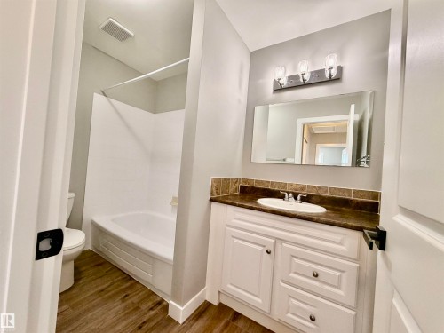 Bathroom featuring wood-finish flooring, a white vanity with an integrated sink and dark countertop, a rectangular mirror, a three-light vanity fixture, and a white bathtub with a shower surround - 11430 139 Avenue Nw, Edmonton, AB - Indoor Photo Showing Bathroom