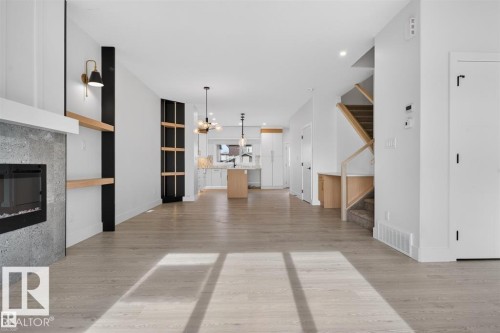 Open-concept living area featuring light wood-finish flooring, a modern fireplace with a grey stone surround, built-in shelving, a kitchen with an island, and a staircase with natural wood accents - 43 River Birch Way, St. Albert, AB - Indoor With Fireplace
