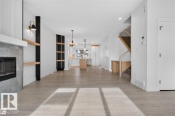 Open-concept living area featuring light wood-finish flooring, a modern fireplace with a grey stone surround, built-in shelving, a kitchen with an island, and a staircase with natural wood accents - 