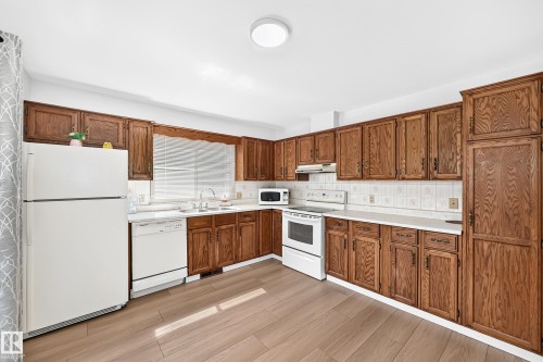 Kitchen featuring wood-finish cabinetry, white appliances, light-toned countertops, tile backsplash, and wood-finish flooring - 6208 11 Ave Nw Nw, Edmonton, AB - Indoor Photo Showing Kitchen With Double Sink