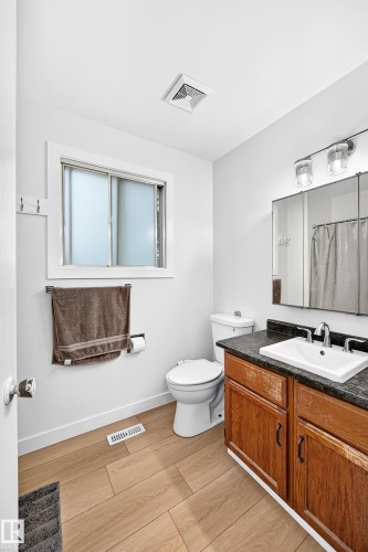 Bathroom featuring wood-finish flooring, a wood vanity with a dark countertop, a white sink, a mirror with integrated lighting, and a frosted window - 6208 11 Ave Nw Nw, Edmonton, AB - Indoor Photo Showing Bathroom