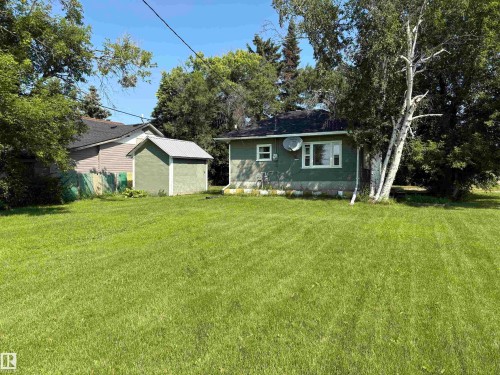 Expansive lawn with mature trees, a detached storage shed with a metal roof, and a single-story residence featuring green siding and white trim windows - 4901 53 Avenue, Elk Point, AB - Outdoor