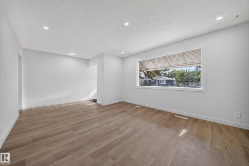Spacious living area featuring wood-finish flooring, a large horizontal window, recessed ceiling lighting, textured ceilings, and light-toned walls - 5204 97A Avenue Nw, Edmonton, AB - Indoor Photo Showing Other Room