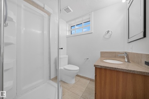 Bathroom featuring a glass-enclosed shower, a wood-finish vanity with a stone countertop, a white toilet, and a window with white trim - 5204 97A Avenue Nw, Edmonton, AB - Indoor Photo Showing Bathroom