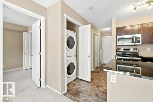 Integrated laundry closet with stacked appliances, featuring a tiled floor, dark wood-finish cabinetry, stainless steel kitchen appliances, and dark countertops - #119 111 Watt Cm Sw, Edmonton, AB - Indoor Photo Showing Laundry Room