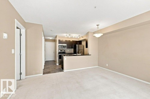 Open-concept living area featuring light-toned carpeting and a kitchen with dark cabinetry, stainless steel appliances, and a black countertop - #119 111 Watt Cm Sw, Edmonton, AB - Indoor