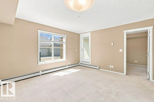 Neutral-toned room featuring a large window with white trim, light beige carpeting, and a white exterior door with vertical siding - #119 111 Watt Cm Sw, Edmonton, AB - Indoor Photo Showing Other Room