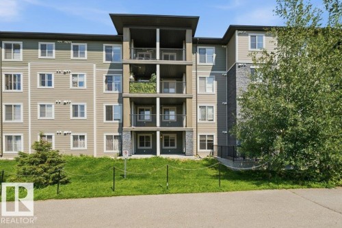Modern multi-story building featuring multiple balconies with black railings, a contrasting exterior of beige and green siding, stone accent panels, and a paved frontage - #119 111 Watt Cm Sw, Edmonton, AB - Outdoor With Facade