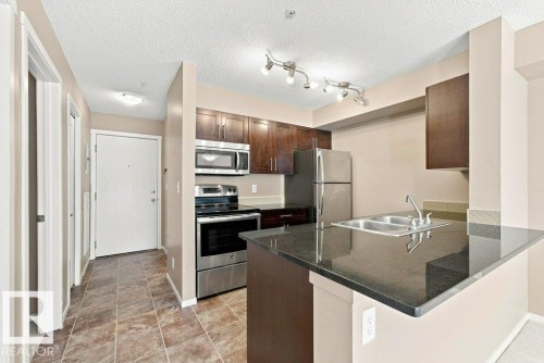 Kitchen featuring dark wood cabinetry, stainless steel appliances, and a black countertop with an integrated double sink - #119 111 Watt Cm Sw, Edmonton, AB - Indoor Photo Showing Kitchen With Double Sink