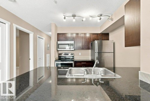 Kitchen featuring dark cabinetry, stainless steel appliances, track lighting, a double basin sink, and a dark stone countertop - #119 111 Watt Cm Sw, Edmonton, AB - Indoor Photo Showing Kitchen With Double Sink