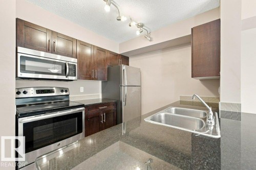 Kitchen featuring dark wood cabinetry, stainless steel appliances, a double basin sink, and dark stone countertops - #119 111 Watt Cm Sw, Edmonton, AB - Indoor Photo Showing Kitchen With Double Sink
