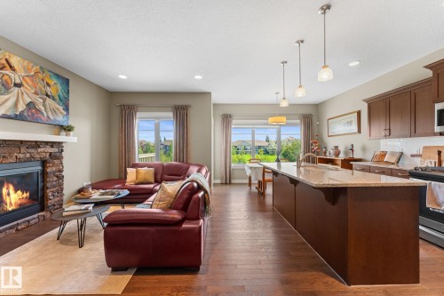 Open-concept living space featuring wood-finish flooring, a stone-clad fireplace with a white mantel, and a kitchen island with a light-colored countertop - 3413 Cameron Heights Cove Nw, Edmonton, AB - Indoor With Fireplace