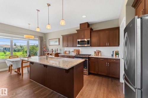 Kitchen featuring a granite-topped island with an undermount sink, rich wood-finish cabinetry, stainless steel appliances, and wood-finish flooring - 3413 Cameron Heights Cove Nw, Edmonton, AB - Indoor Photo Showing Kitchen With Upgraded Kitchen