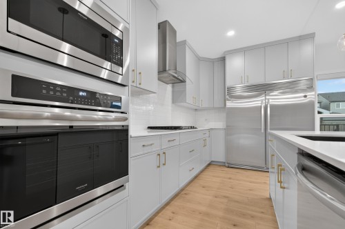 Modern kitchen featuring light wood-finish flooring, white shaker-style cabinetry with gold-tone hardware, stainless steel appliances including a built-in oven and microwave, and a white subway tile backsplash - 1534 Plum Circle Circle Sw, Edmonton, AB - Indoor Photo Showing Kitchen
