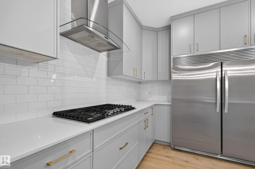 Contemporary kitchen featuring light gray cabinetry with gold-finish hardware, a built-in stainless steel refrigerator, white subway tile backsplash, white countertops, and wood-finish flooring - 1534 Plum Circle Circle Sw, Edmonton, AB - Indoor Photo Showing Kitchen