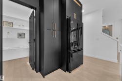 Integrated black French door refrigerator, matte black cabinetry with brass hardware, light wood-finish flooring, and pantry with white shelving - 