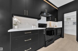Modern kitchen featuring black cabinetry with gold-tone hardware, white countertops, a contrasting white backsplash, and wood-finish flooring - 