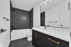 Bathroom featuring a modern vanity with a white countertop, dark cabinetry with gold-toned hardware, a rectangular mirror, and a bathtub with dark speckled tile surround - 