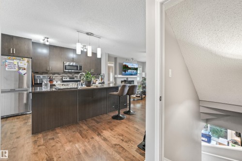 Modern kitchen featuring wood-finish flooring, dark wood cabinetry, a stainless steel refrigerator, and an island with built-in seating - #40 1030 Chappelle Bv Sw, Edmonton, AB - Indoor Photo Showing Kitchen With Upgraded Kitchen
