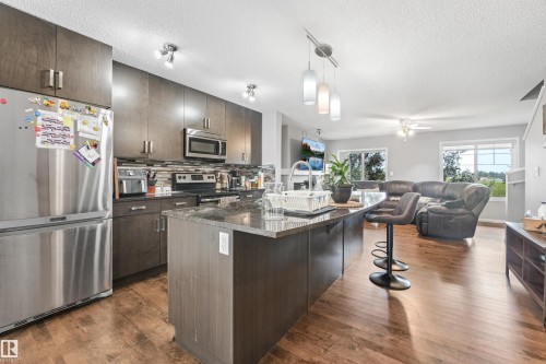 Open-concept kitchen featuring dark wood-finish cabinetry, stainless steel appliances, a tile backsplash, and a central island with a stone countertop and integrated sink - #40 1030 Chappelle Bv Sw, Edmonton, AB - Indoor Photo Showing Kitchen With Stainless Steel Kitchen With Upgraded Kitchen