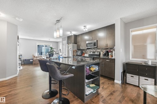 Kitchen featuring dark wood-finish cabinetry, stainless steel appliances, a dark granite-style island with built-in shelving, and wood-finish flooring - #40 1030 Chappelle Bv Sw, Edmonton, AB - Indoor