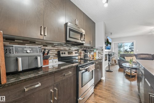 Contemporary kitchen featuring dark wood-finish cabinetry, stainless steel appliances, a multi-tone tile backsplash, and dark stone countertops - #40 1030 Chappelle Bv Sw, Edmonton, AB - Indoor Photo Showing Kitchen With Stainless Steel Kitchen