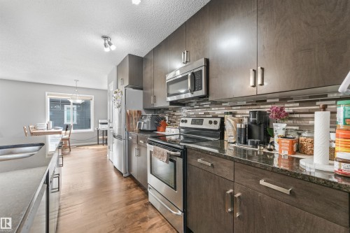 Contemporary kitchen featuring dark wood-finish cabinetry, stainless steel appliances, dark stone countertops, and a mosaic tile backsplash - #40 1030 Chappelle Bv Sw, Edmonton, AB - Indoor Photo Showing Kitchen With Stainless Steel Kitchen With Upgraded Kitchen