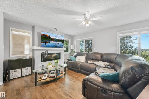 Living area featuring wood-finish flooring, a contemporary fireplace with a white mantle, a ceiling fan with integrated lighting, and multiple windows with horizontal blinds - #40 1030 Chappelle Bv Sw, Edmonton, AB - Indoor Photo Showing Living Room With Fireplace