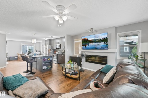 Open-concept living area featuring wood-finish flooring, a contemporary electric fireplace, a ceiling fan with integrated lighting, a kitchen island with seating, and stainless steel kitchen appliances - #40 1030 Chappelle Bv Sw, Edmonton, AB - Indoor Photo Showing Living Room With Fireplace
