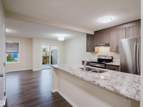 Open-concept kitchen and dining area featuring a breakfast bar with dual sinks, speckled countertop, wood-finish cabinetry, and stainless steel appliances - 5407 14 Avenue Sw, Edmonton, AB - Indoor Photo Showing Kitchen With Stainless Steel Kitchen With Double Sink With Upgraded Kitchen