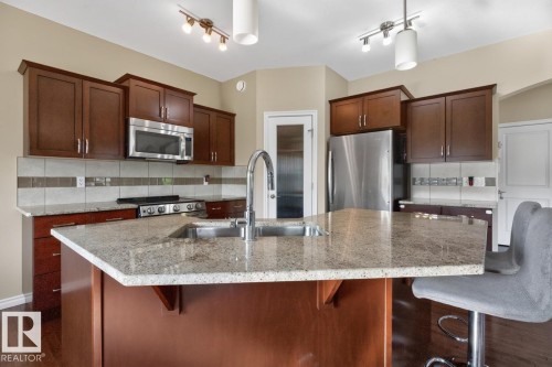Kitchen featuring wood-finish cabinetry, stainless steel appliances, a multi-tone tile backsplash, a large island with an integrated double sink, and stone-look countertops - 109 Selkirk Place, Leduc, AB - Indoor Photo Showing Kitchen With Double Sink