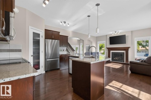 Open-concept kitchen and living area featuring wood-finish flooring, a kitchen island with pendant lighting, stone countertops, stainless steel appliances, and a fireplace with a wood mantle - 109 Selkirk Place, Leduc, AB - Indoor Photo Showing Kitchen With Fireplace
