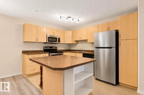 Kitchen featuring light wood-finish cabinetry, stainless steel appliances, and an island with open shelving - #203 17407 99 Av Nw, Edmonton, AB - Indoor Photo Showing Kitchen With Double Sink