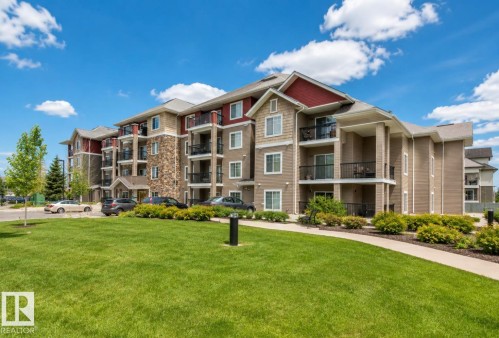 Multi-story residential building featuring a stone and siding exterior, private balconies with black railings, and a well-maintained lawn with mature landscaping - #203 17407 99 Av Nw, Edmonton, AB - Outdoor With Facade