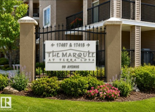 Exterior entry featuring a custom sign, wrought iron fence, stucco pillars, and established landscaping with green shrubs and flowering plants - #203 17407 99 Av Nw, Edmonton, AB - Outdoor