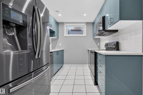 Galley kitchen featuring stainless steel appliances, blue high-gloss cabinetry, white countertop surfaces, a textured white tile backsplash, and white square floor tiles - 12136 135A Avenue Nw, Edmonton, AB - Indoor Photo Showing Kitchen