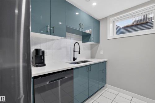 Kitchenette featuring glossy teal cabinetry, white countertop, stainless steel sink with matte black faucet, integrated dishwasher, and tiled flooring - 12136 135A Avenue Nw, Edmonton, AB - Indoor Photo Showing Kitchen With Double Sink