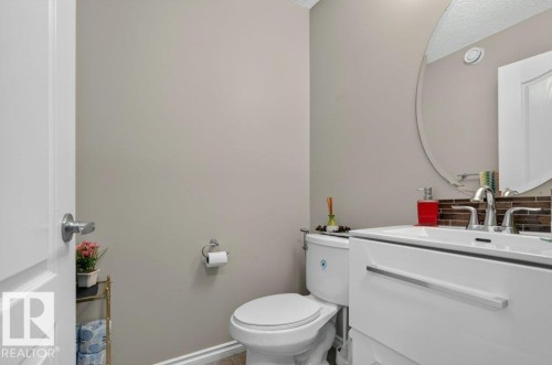 Contemporary powder room featuring a white single-sink vanity with chrome hardware, an oval frameless mirror, and a brown mosaic tile backsplash - 692 Secord Boulevard Nw, Edmonton, AB - Indoor Photo Showing Bathroom