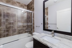 Bathroom featuring a white bathtub with sliding glass doors, brown tiled shower surround, dark wood-finish vanity with an integrated rectangular sink, stone countertop, and a large framed mirror - 