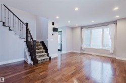 Spacious room featuring wood-finish flooring, a prominent staircase with dark wood newel post and wrought iron balusters, and a large multi-pane window - 