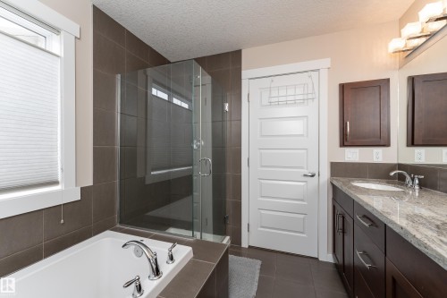 Bathroom featuring a built-in soaking tub with chrome fixtures, a glass-enclosed shower, a double vanity with a stone countertop and integrated sinks, brown tile flooring, and a white panel door - 2338 Frezenberg Avenue Nw, Edmonton, AB - Indoor Photo Showing Bathroom