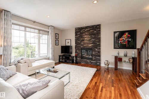Living area featuring rich wood-finish flooring, a stacked stone fireplace, large windows, recessed lighting, and an open staircase with wood treads and metal spindles - 2338 Frezenberg Avenue Nw, Edmonton, AB - Indoor Photo Showing Living Room With Fireplace