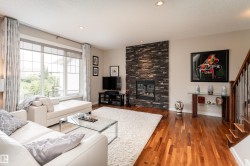 Living area featuring rich wood-finish flooring, a stacked stone fireplace, large windows, recessed lighting, and an open staircase with wood treads and metal spindles - 