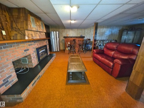 Recreation room featuring a brick fireplace with a black hearth, textured wood-finish wall panels, a built-in bar area, and recessed ceiling lighting - 5201 54 Street, Elk Point, AB - Indoor With Fireplace