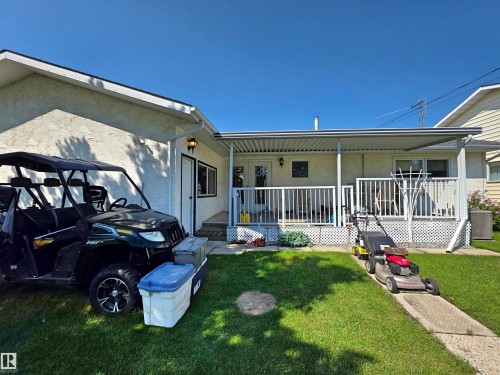 Rear exterior featuring a covered patio with white railing, stucco siding, a maintained lawn, and a concrete pathway - 5201 54 Street, Elk Point, AB - Outdoor