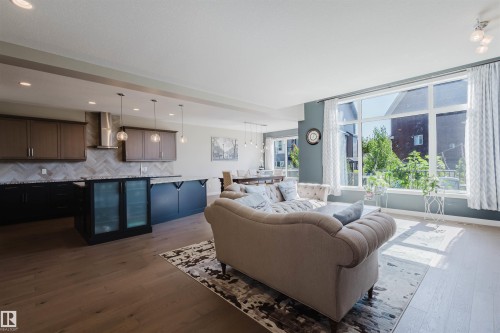 Open-concept living space featuring wood-finish flooring, a kitchen island with integrated beverage coolers, and a herringbone tile backsplash - 4125 Kennedy Green Sw, Edmonton, AB - Indoor
