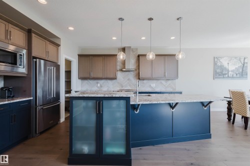 Spacious kitchen featuring an expansive center island with seating, pendant lighting, stainless steel appliances, a herringbone tile backsplash, and wood-finish flooring - 4125 Kennedy Green Sw, Edmonton, AB - Indoor Photo Showing Kitchen With Upgraded Kitchen